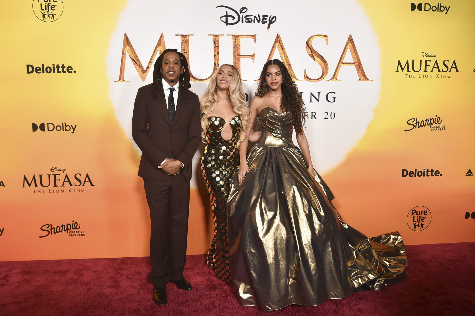JAY-Z, Beyoncé y su hija Blue Ivy Carter posan en la alfombra roja durante el estreno de la película de Disney Mufasa: The Lion King. JAY-Z viste un traje oscuro, Beyoncé lleva un vestido negro con detalles dorados y Blue Ivy luce un vestido largo de color dorado metálico.